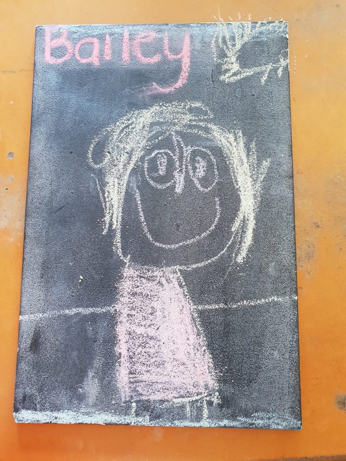 A chalkboard self-portrait in a pink dress, drawn by a Tiny Town child and signed Bailey