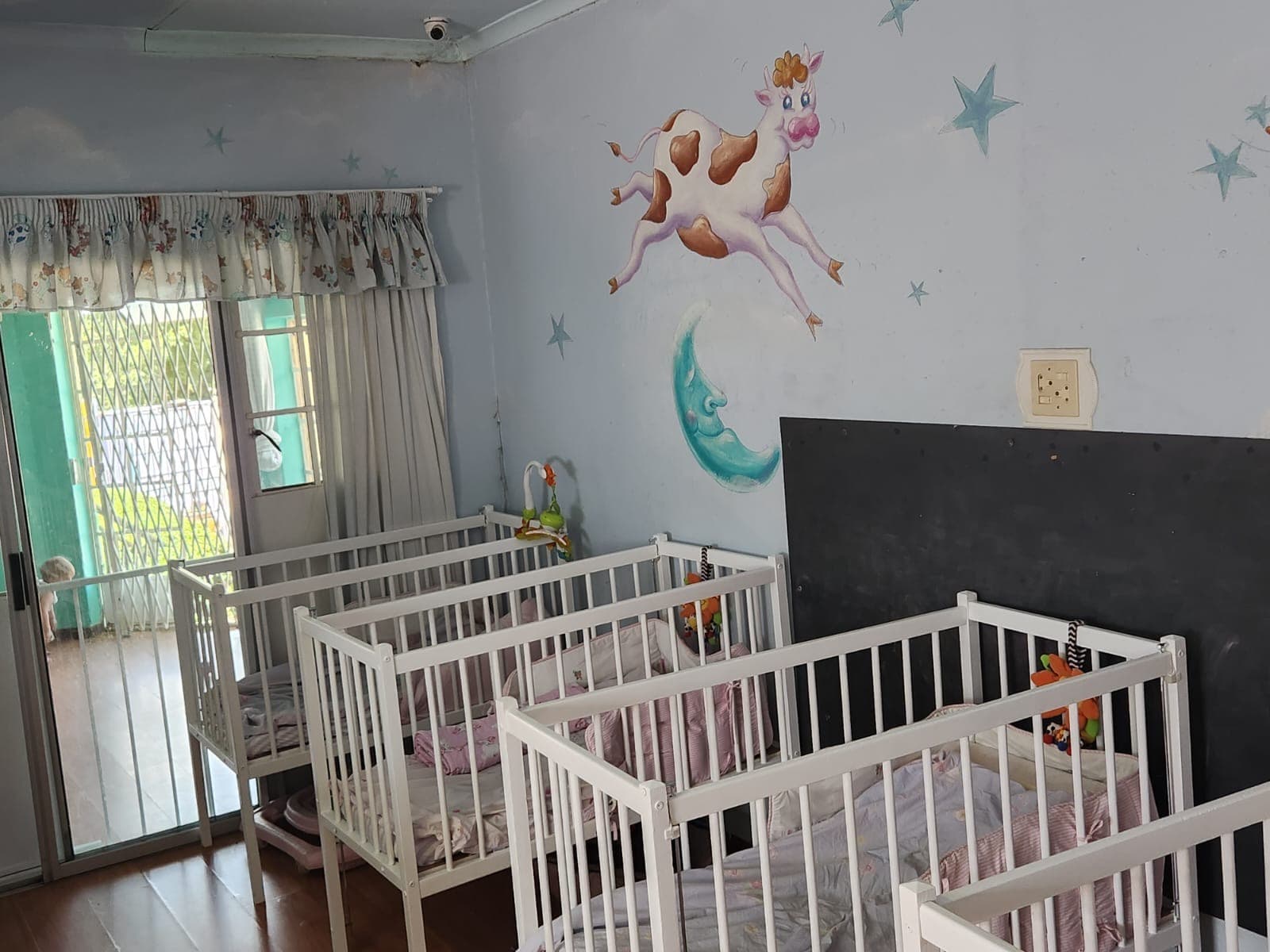The Tiny Town cot room, a row of white cots beneath a cow-jumping-over-the-moon mural