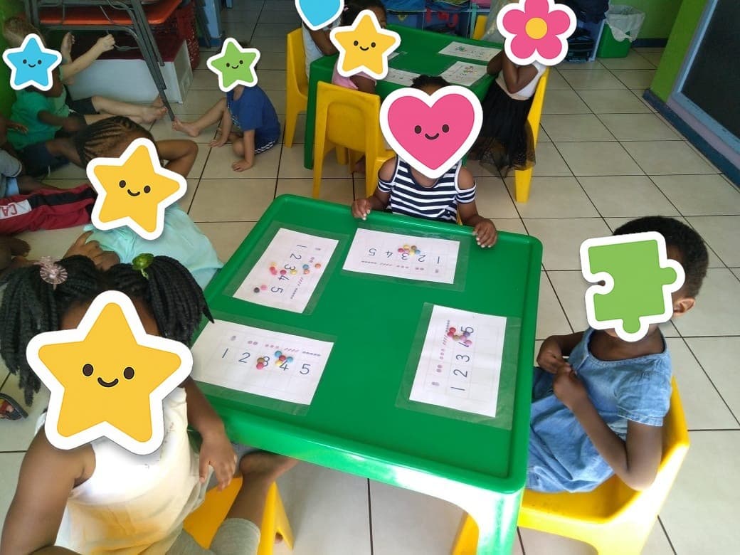 A group of Tiny Town children working at the green tables, holding up their worksheets, with paper stickers covering their faces
